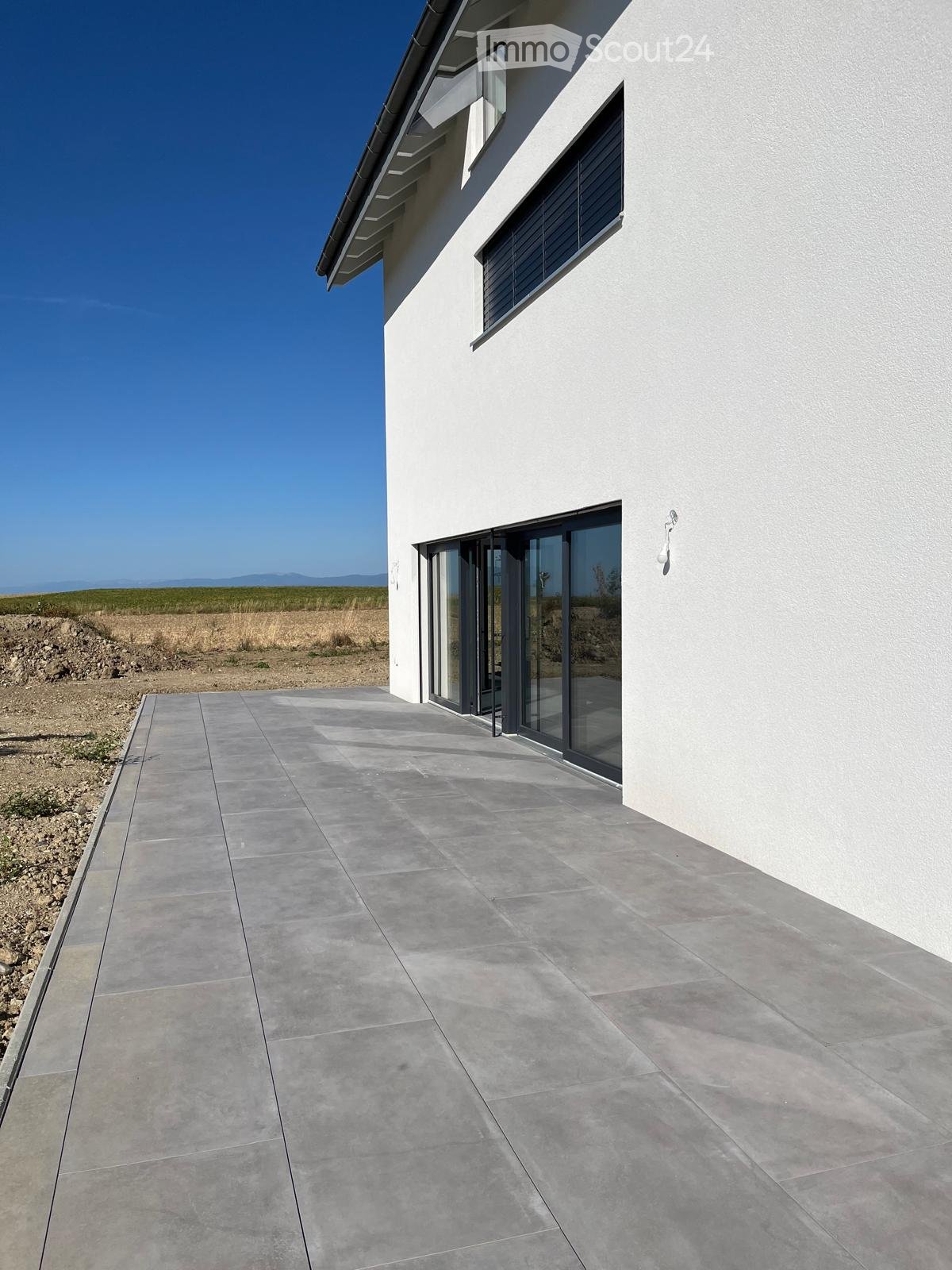 Modern white building with large glass sliding doors, concrete tiled pathway, and open landscape view.