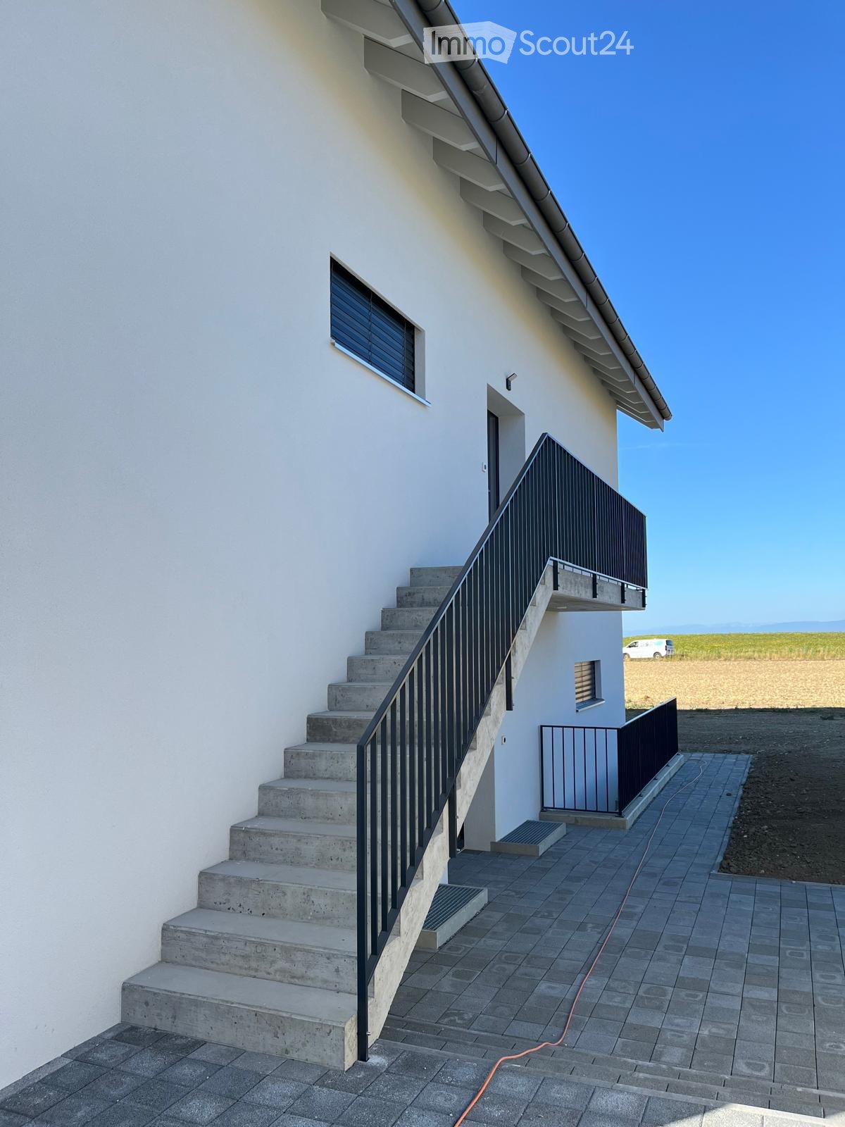 Exterior view of a modern two-story house with white walls, concrete steps leading to a raised ground floor, and a black metal railing.