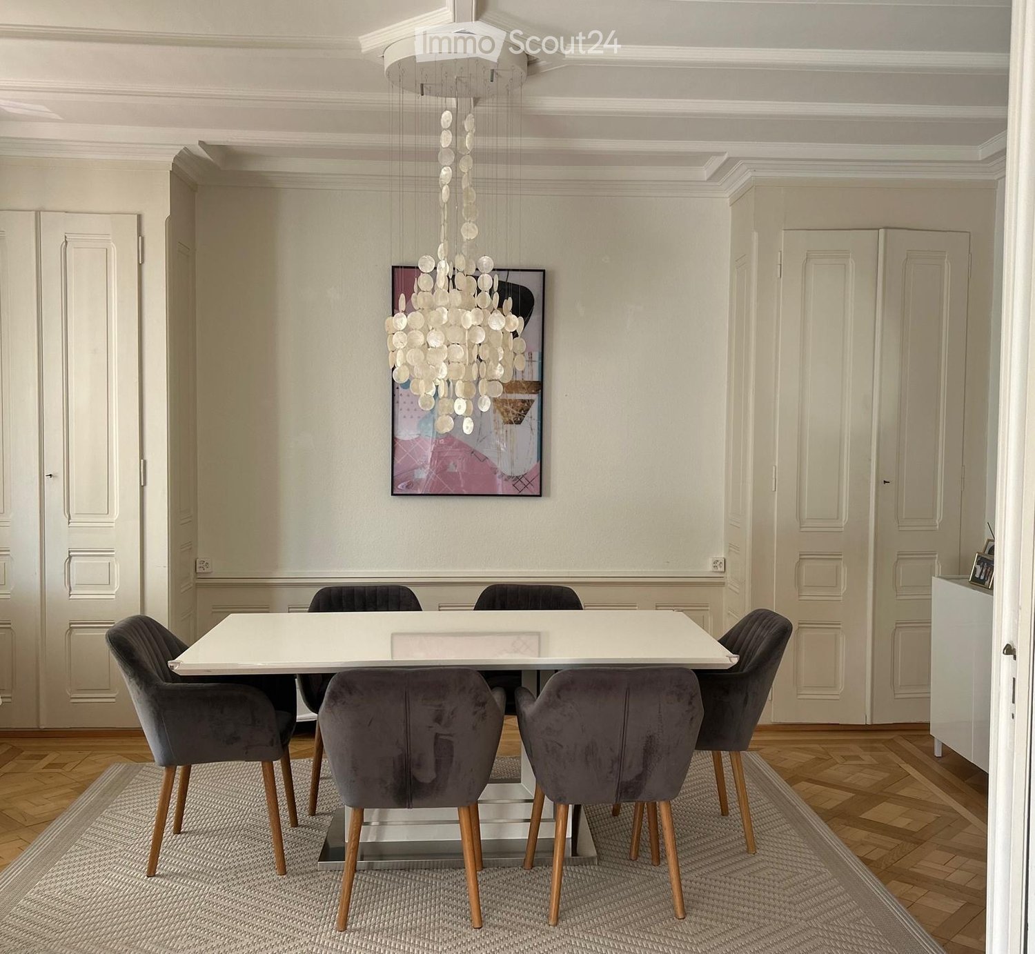 Elegant dining room with a white dining table, six dark grey chairs, a modern chandelier, and a painting on the wall.