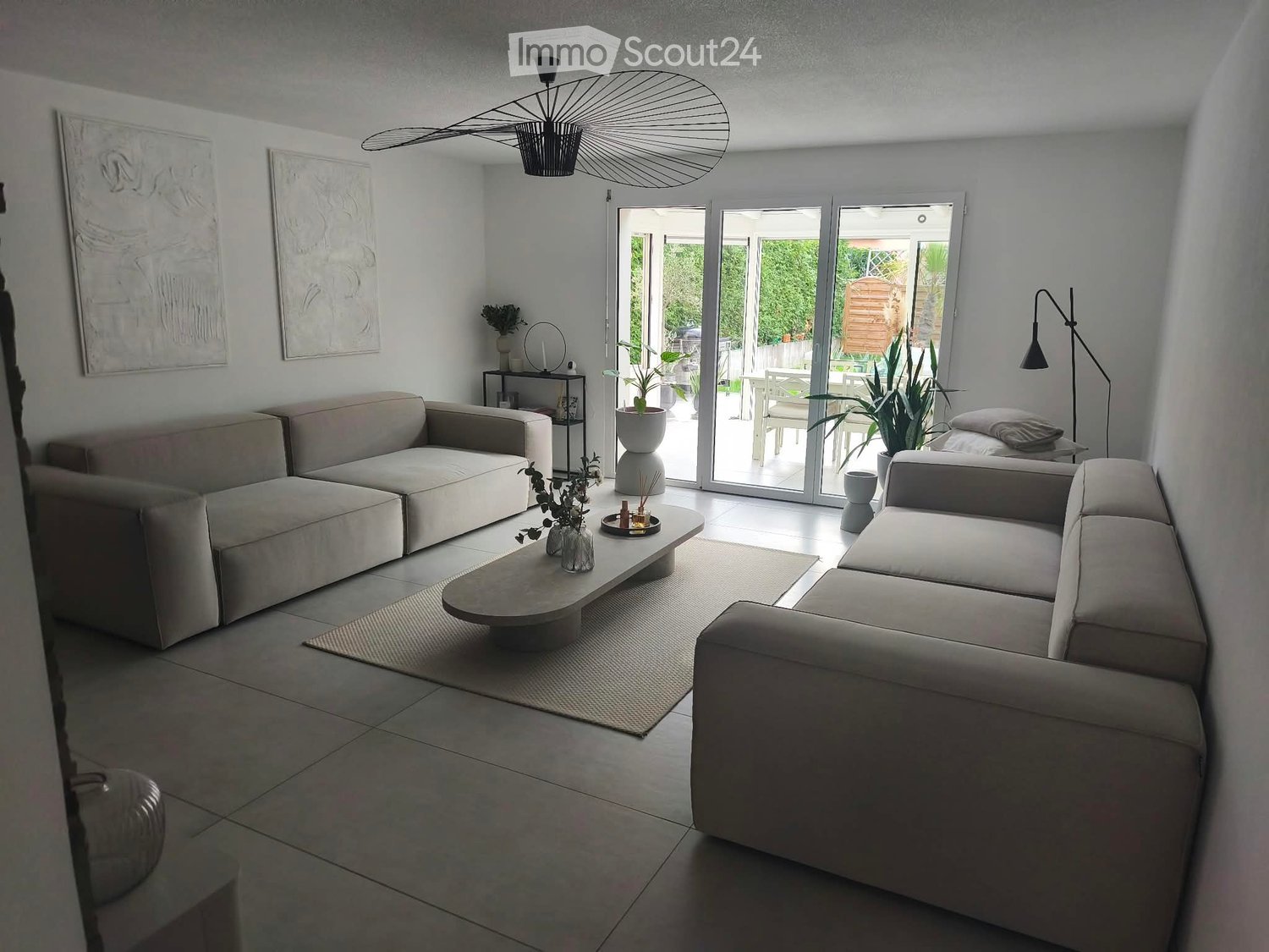 Modern living room with light-colored sofas, a coffee table, floor lamp, and large windows offering a view of the garden.