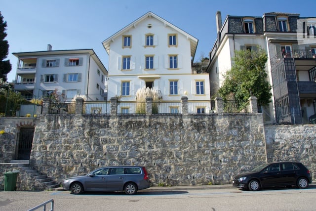 A multi-story residential building with white walls and yellow trim, situated on a raised stone foundation with a small staircase leading to the entrance. Two cars are parked in front of the building.