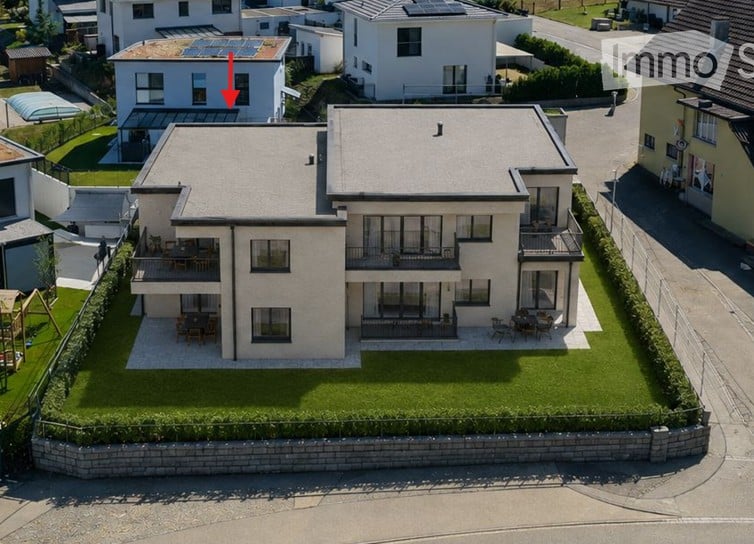 Aerial view of a modern two-story house with a flat roof, surrounded by a well-maintained lawn and neighboring houses. The house features large windows, balconies, and outdoor seating areas.