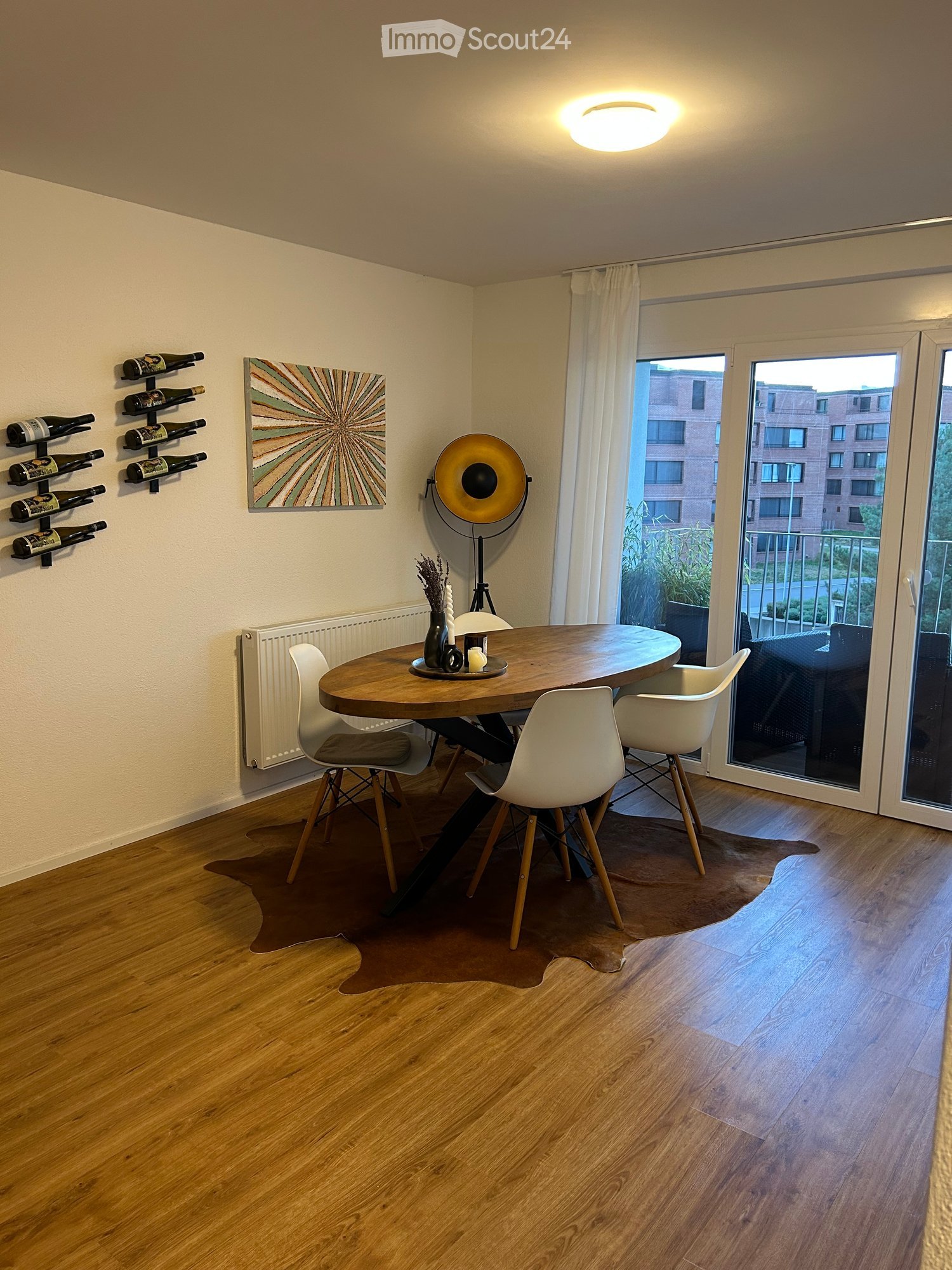Dining area with a round wooden table, four white chairs, a wine rack, a floor lamp, and a decorative painting on the wall.