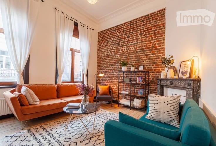 Living room with exposed brick wall, large window with white curtains, orange sofa, teal armchair, coffee table, bookshelf, fireplace, and decorative items.