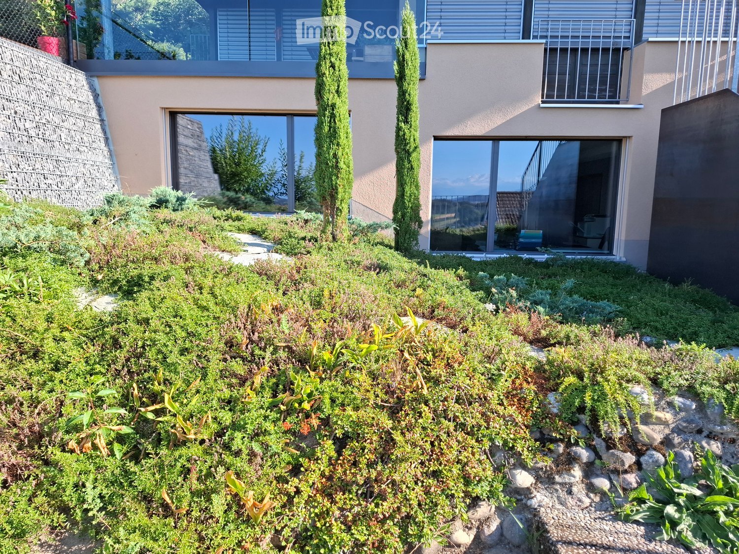Garden with a raised ground floor, featuring a variety of plants, shrubs, and a pathway leading to the house. The house has large windows and a balcony with a metal railing.