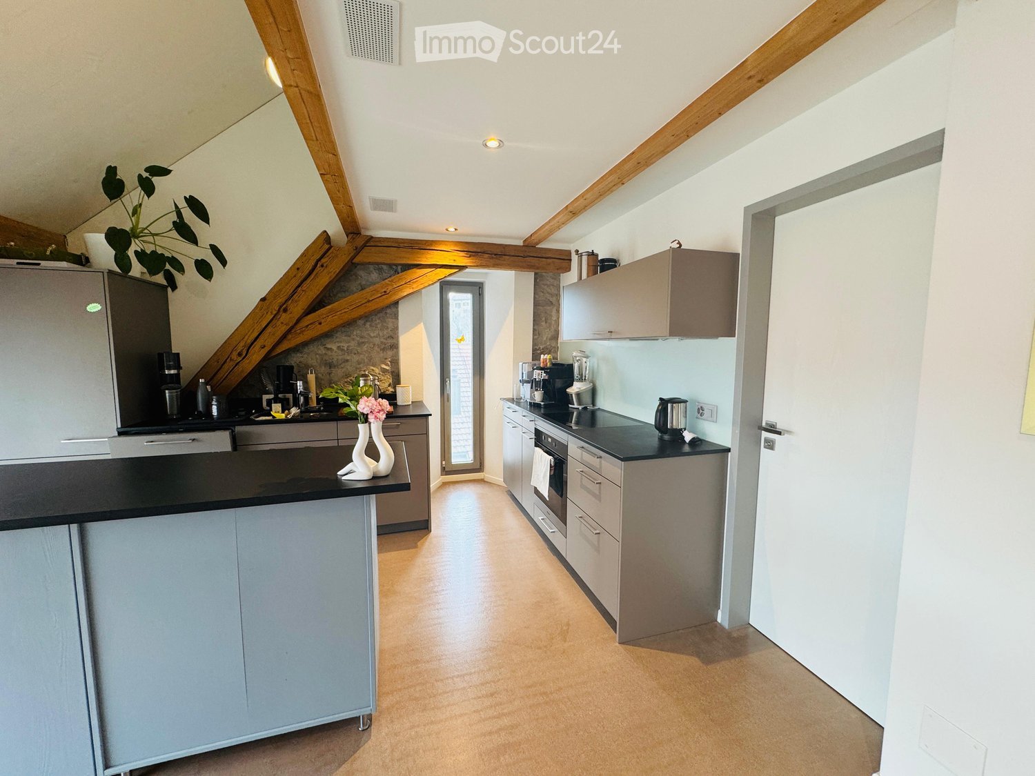 Modern kitchen with light grey cabinets, black countertops, stainless steel appliances, and wooden beams on the ceiling. Features a central island with a vase and plant, and a coffee maker on the counter.