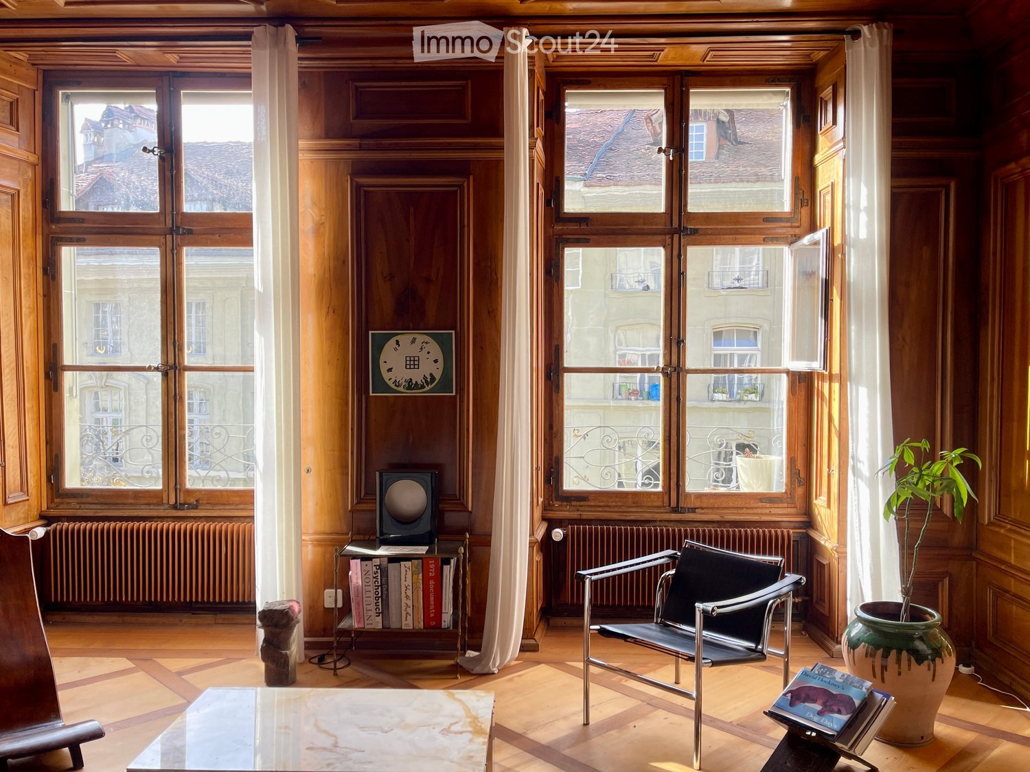 A well-lit room with wooden paneling, large windows, a modern chair, a glass coffee table, a bookshelf with books, a potted plant, and a speaker on a small table.