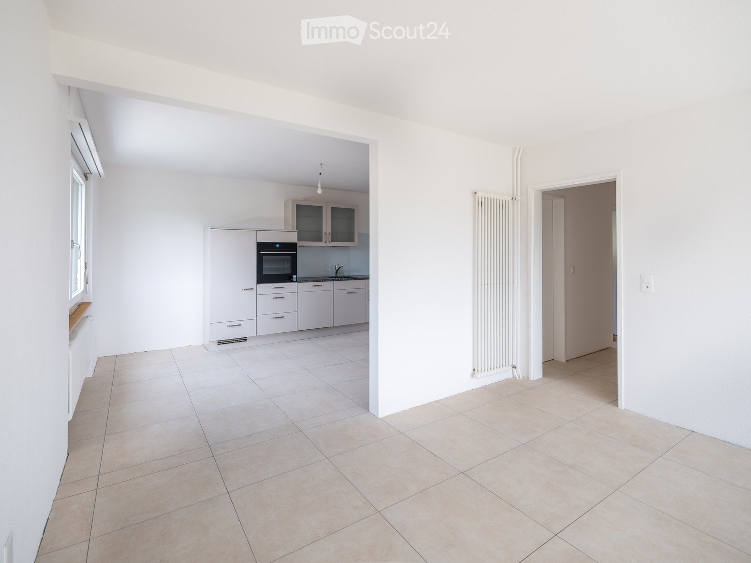 Empty living space with tiled floor, white walls, and a small kitchen area featuring cabinets and an oven. Radiators are visible on the walls.