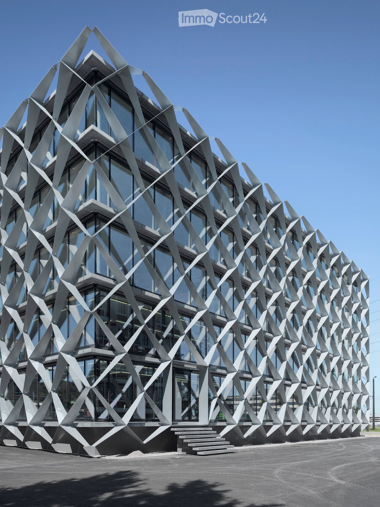 Modern building with a geometric, angular metal facade and large glass windows, featuring a raised ground floor and steps leading to the entrance.