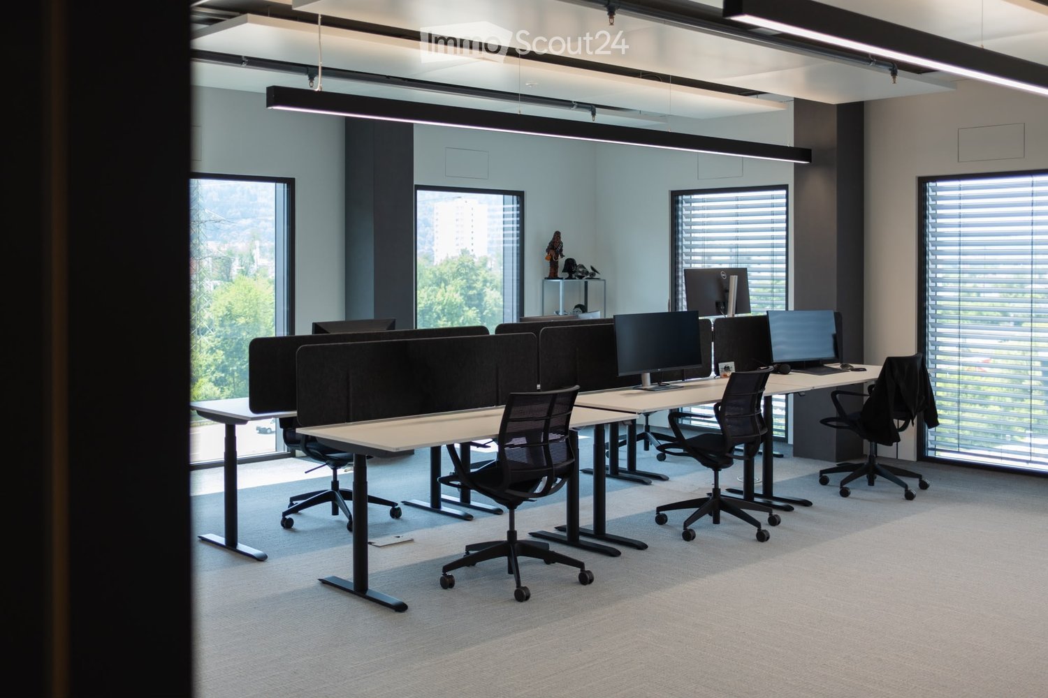 Modern office space with multiple desks, ergonomic chairs, and computer screens arranged in rows. Large windows provide natural light, and the room features a clean, minimalist design with grey tones.