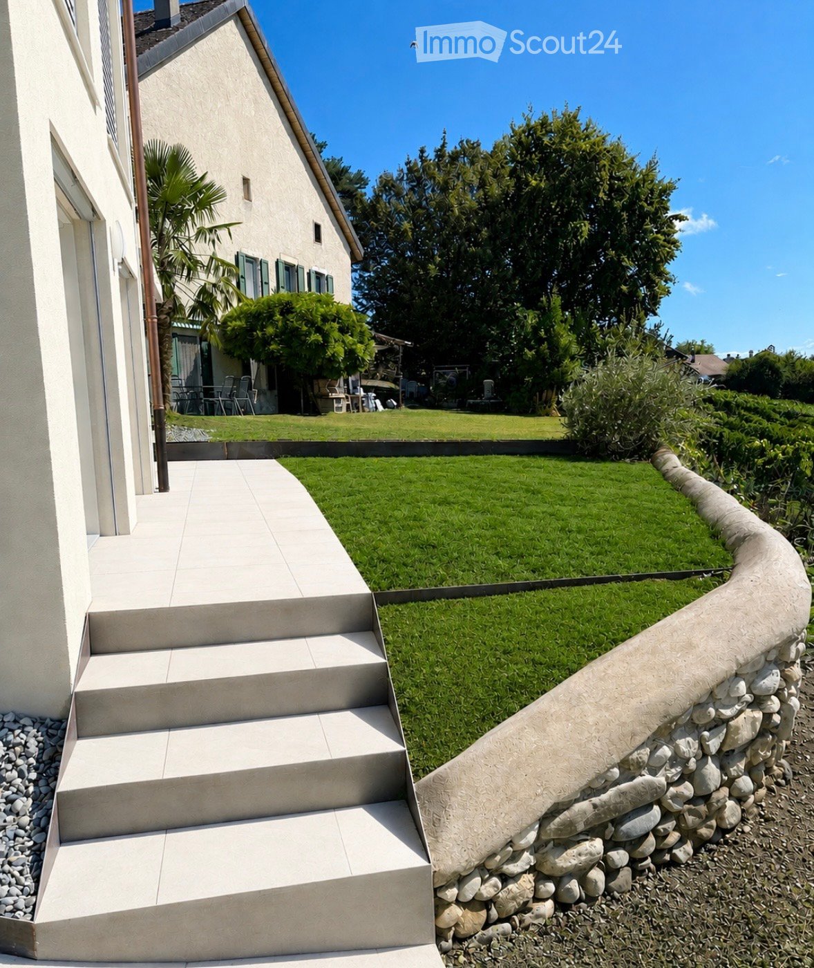 A well-maintained garden with a curved stone wall, steps leading up to a house, and lush greenery.