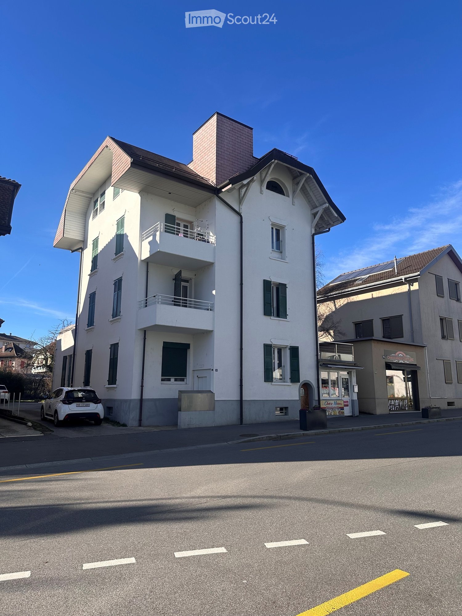 A three-story residential building with white walls, green shutters, and balconies. The building features a chimney and is situated on a street with a parked car and neighboring buildings.