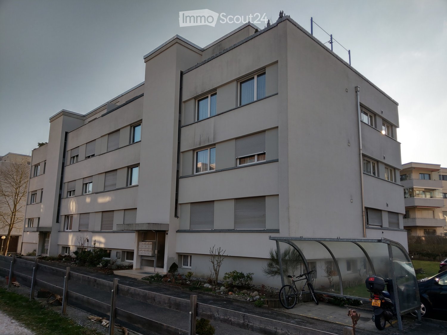 Modern multi-story residential building with grey facade, multiple windows, and balconies. Small landscaped area in front with a bicycle and scooter parked near a bus stop shelter.
