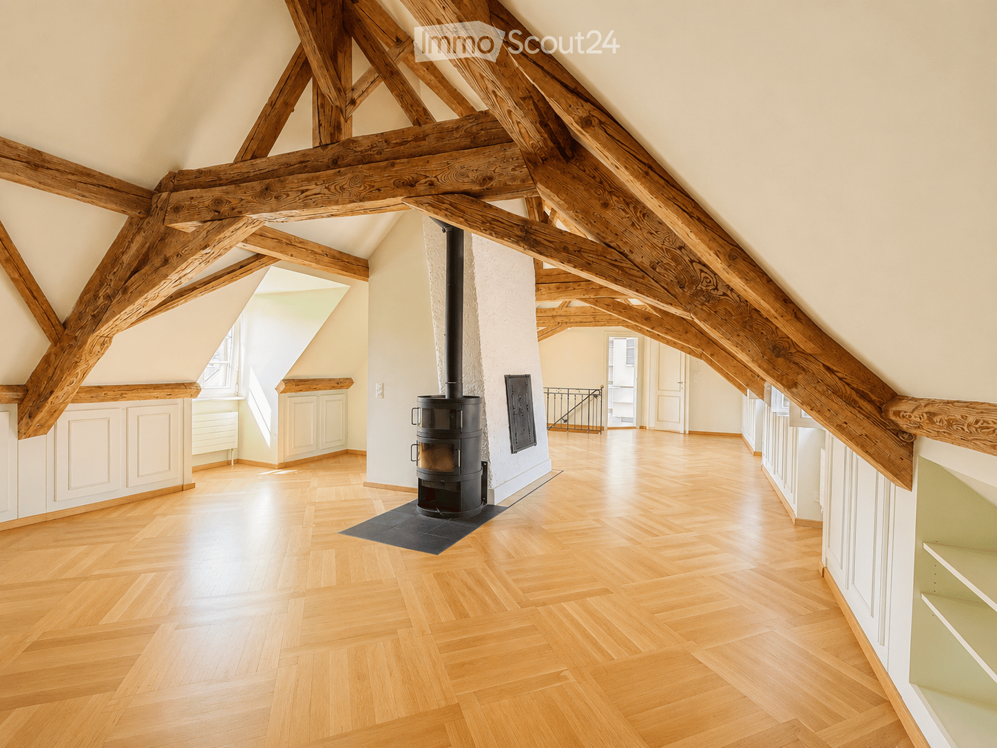 An empty attic space with exposed wooden beams, wooden flooring, and a central wood-burning stove. The room has large windows and white walls.