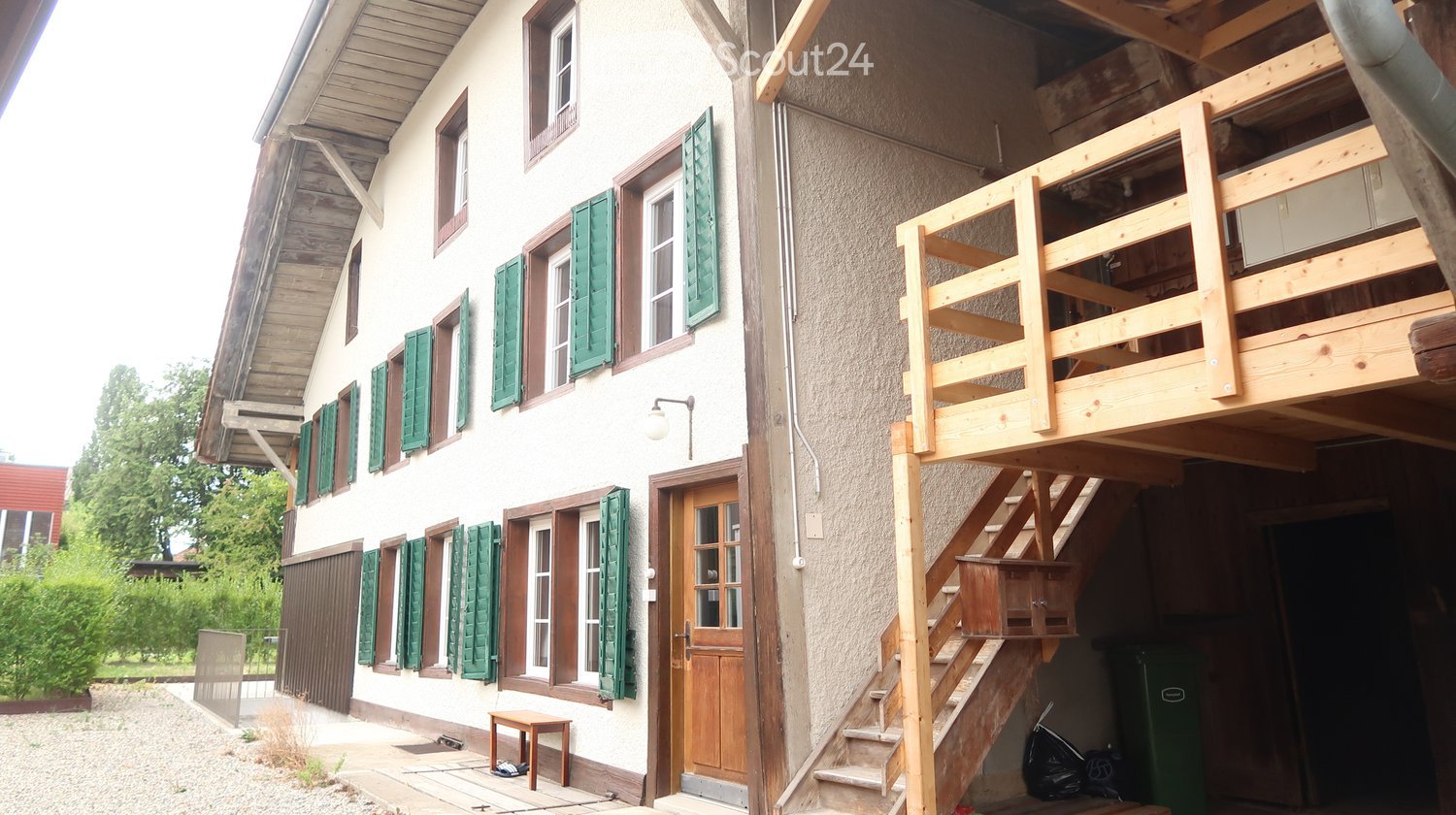 Two-story house with white walls, green shutters, wooden balcony, and wooden staircase leading to the balcony.