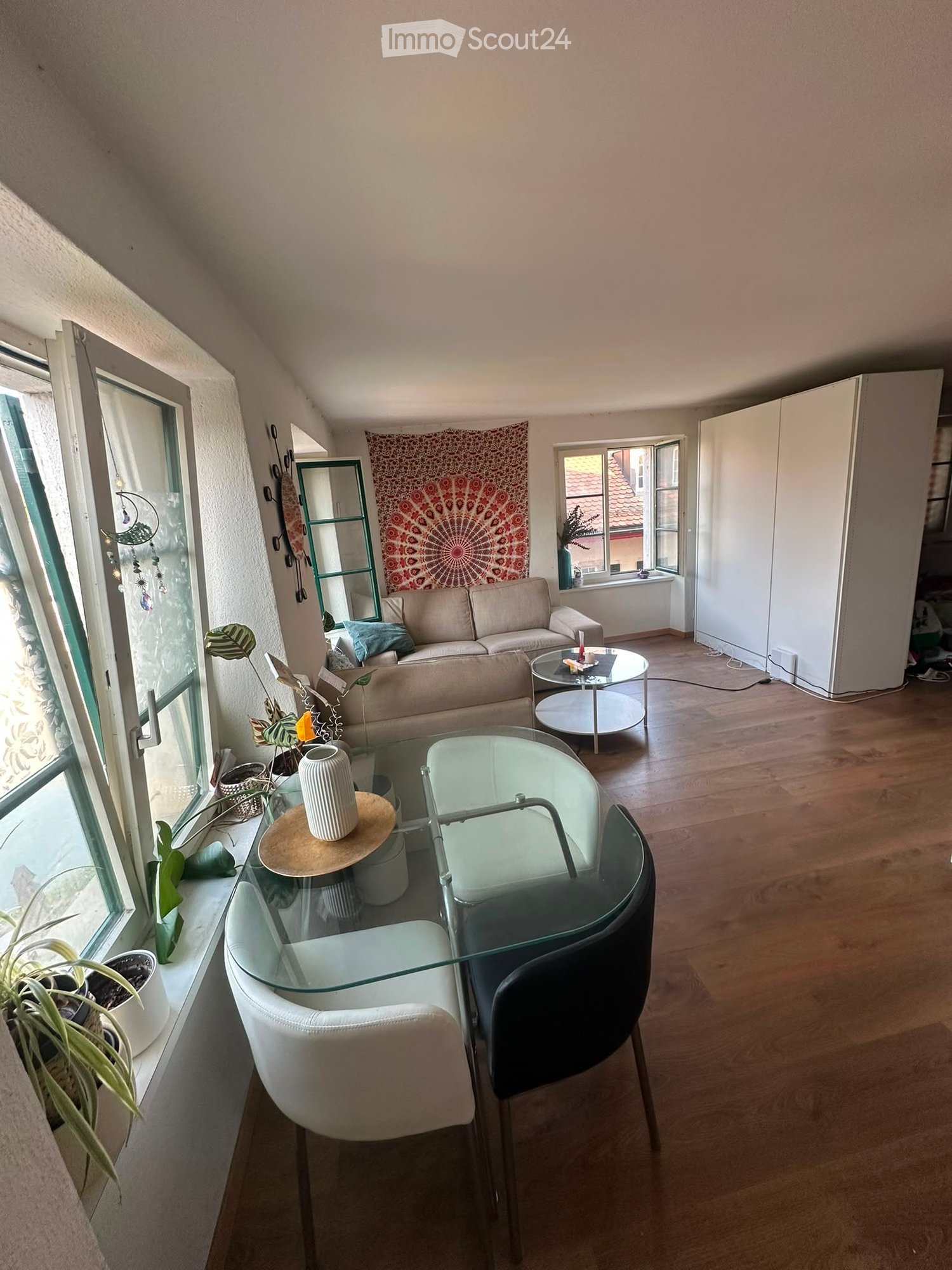 Bright living room with wooden flooring, large windows, a sofa, coffee table, glass dining table with chairs, and decorative vase.