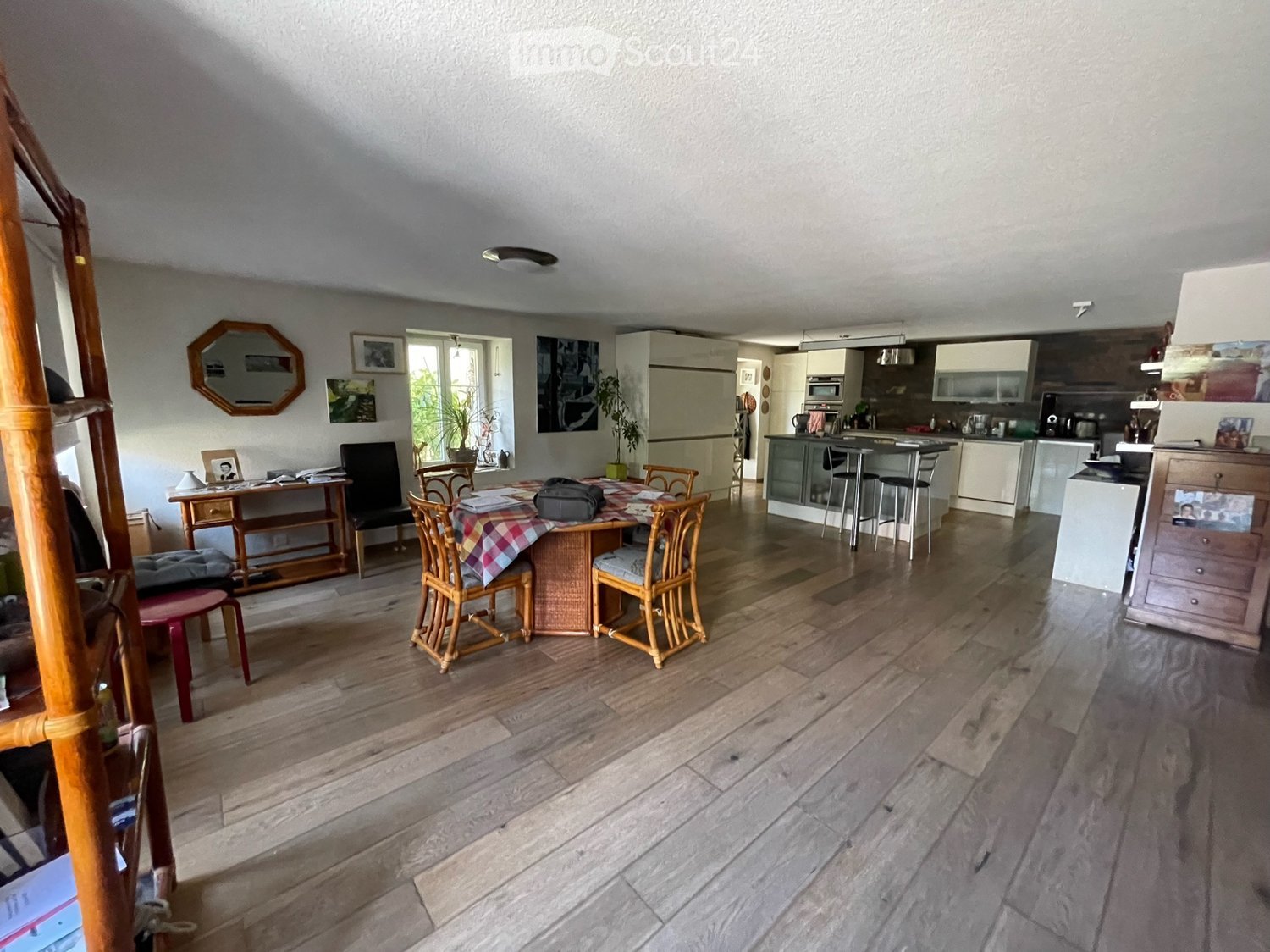 Open-plan living and dining area with wooden flooring, a dining table with chairs, a small cabinet, and a modern kitchen area with appliances.