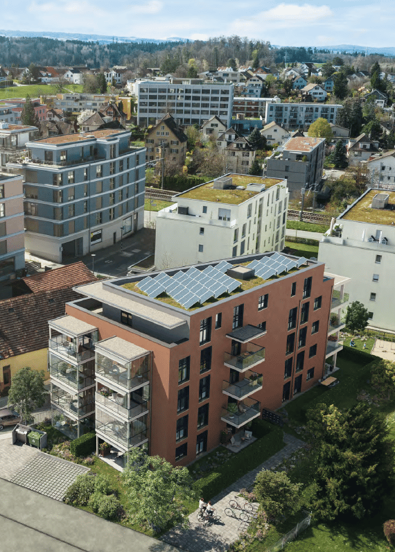 Aerial view of a modern residential complex with multiple buildings, green roofs, solar panels, and surrounding greenery.