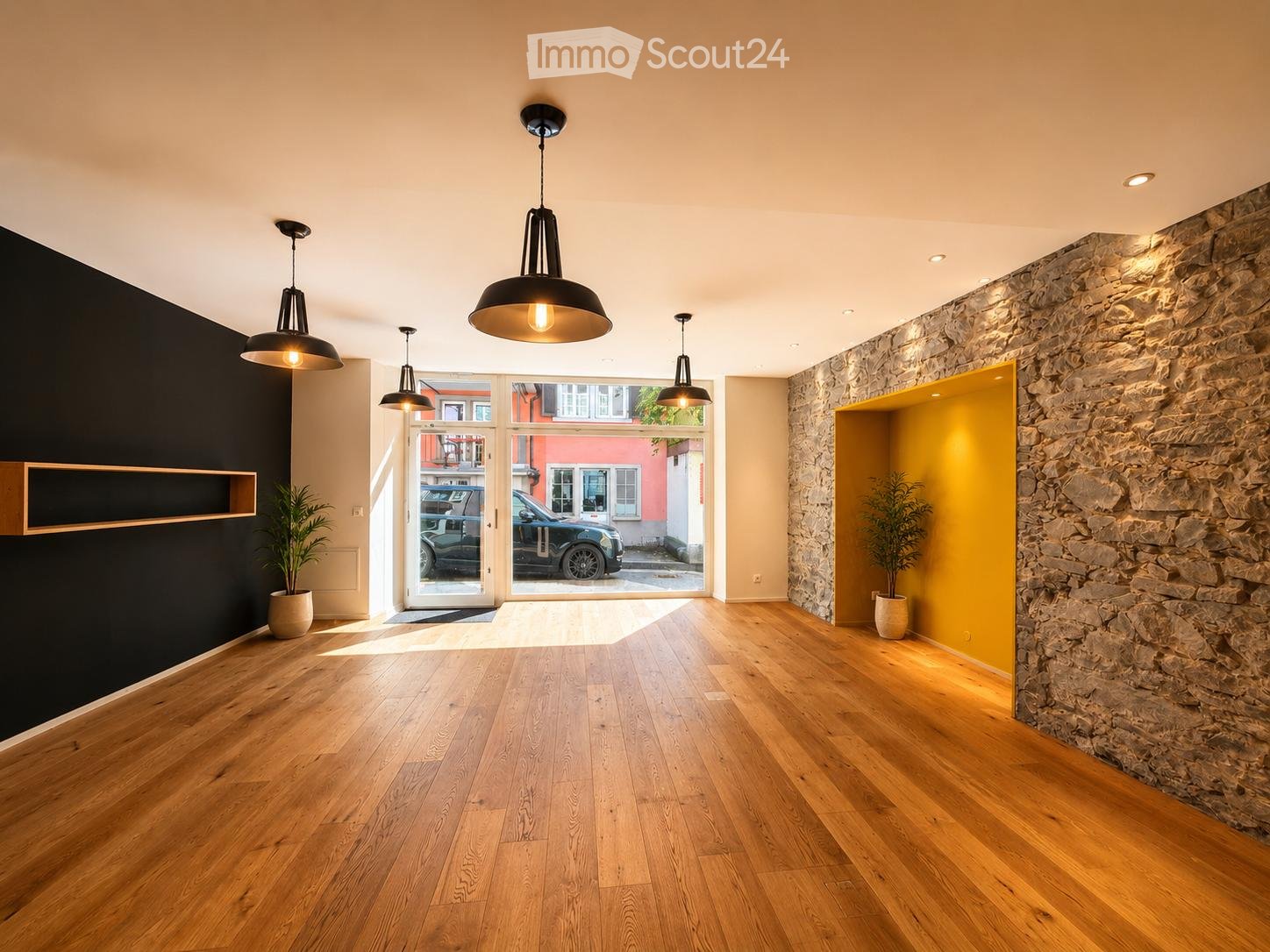 Empty room with wooden flooring, large windows, and modern pendant lights. Features a stone wall and a yellow accent wall.