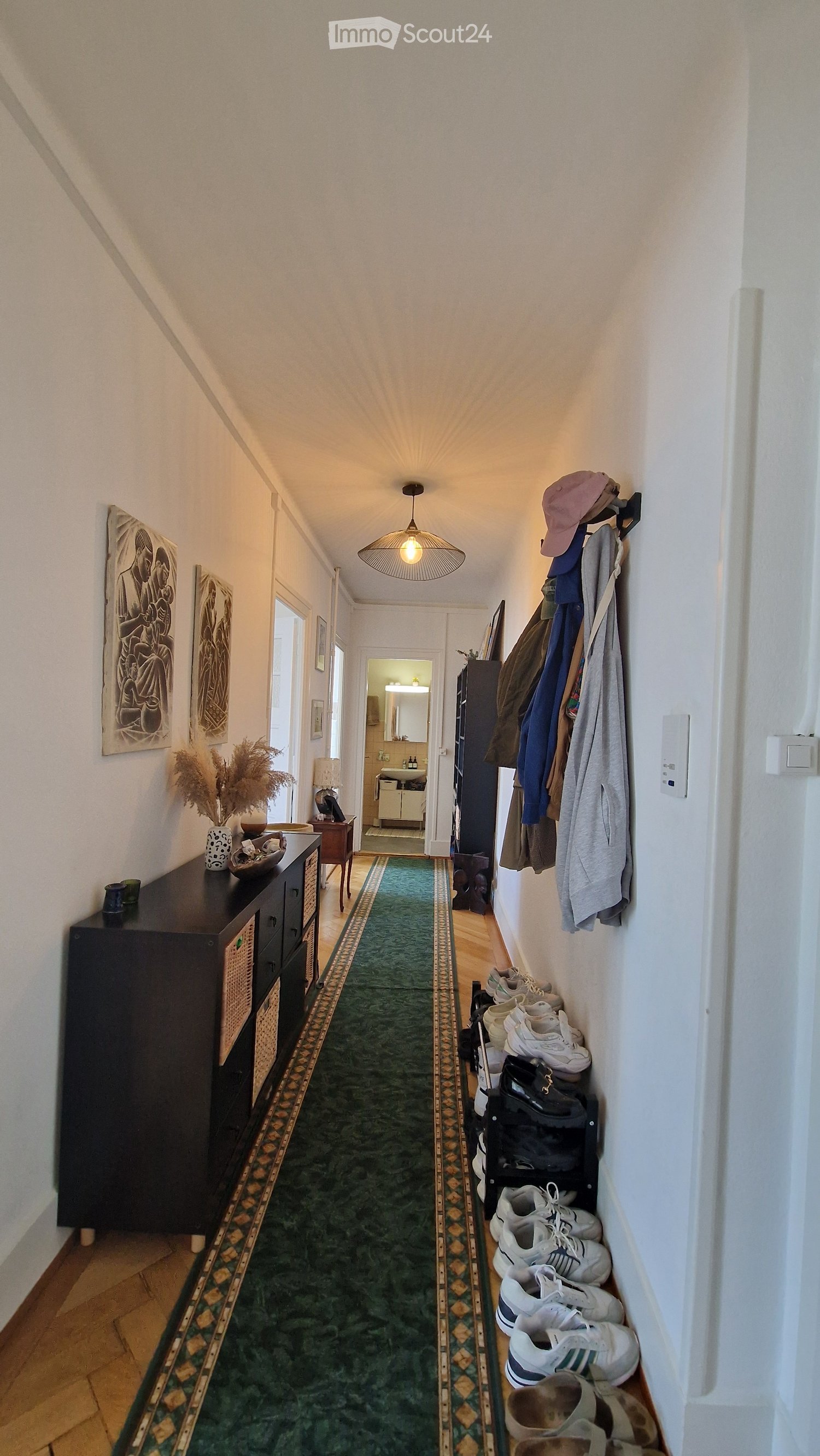 A well-lit hallway with a green runner, wooden flooring, and various items including a coat rack with jackets, shoes, and a small table with decorative items.
