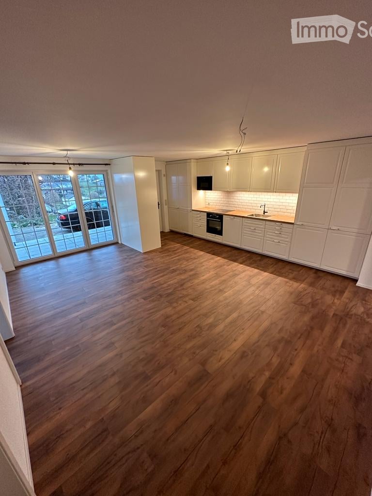 Open-plan space with wooden flooring, white kitchen cabinets, and a large glass sliding door leading to an outdoor area.