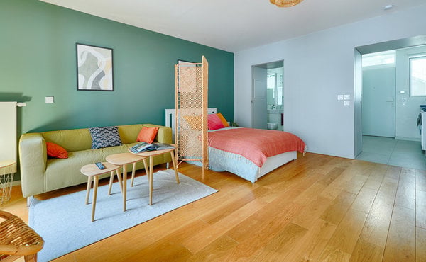A modern, open-concept living and sleeping area with a green accent wall, a light green sofa with colorful pillows, a wooden bed with a pink and orange bedspread, a small wooden table, and a wicker chair. The room features hardwood flooring and a large wi