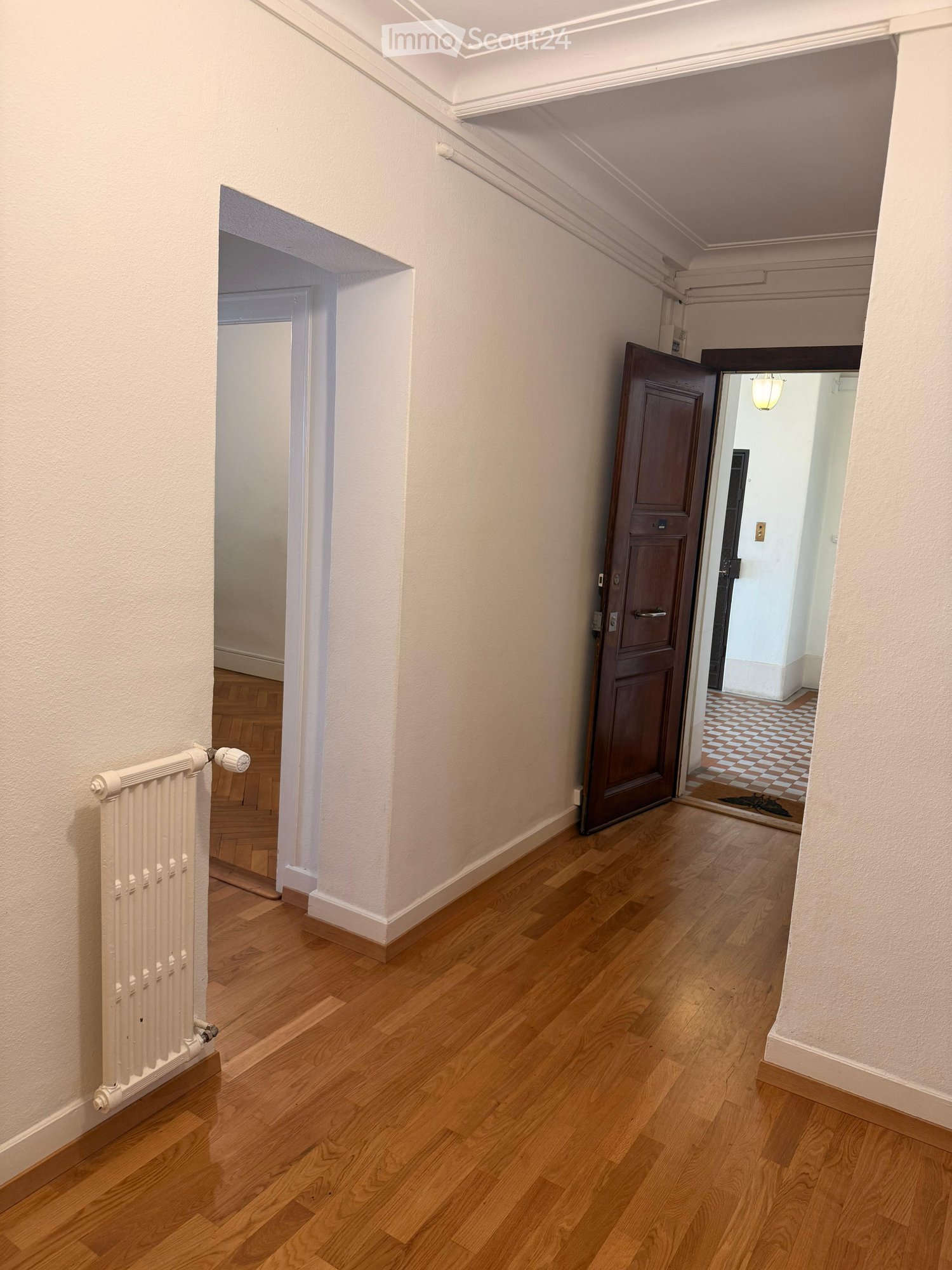 A well-lit hallway with wooden flooring, white walls, and a radiator on the left side. Two doors are visible, one leading to another room with a checkered floor.