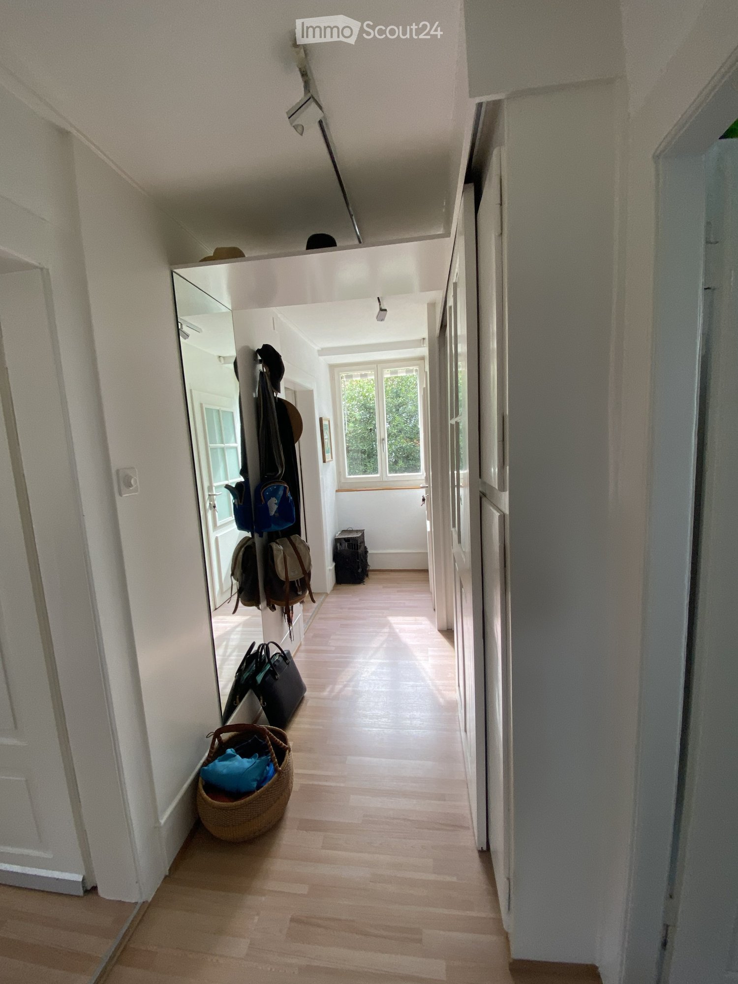 A well-lit hallway with wooden flooring, white walls, and a coat rack holding various bags and hats. A wicker basket with items is placed on the floor near the coat rack.