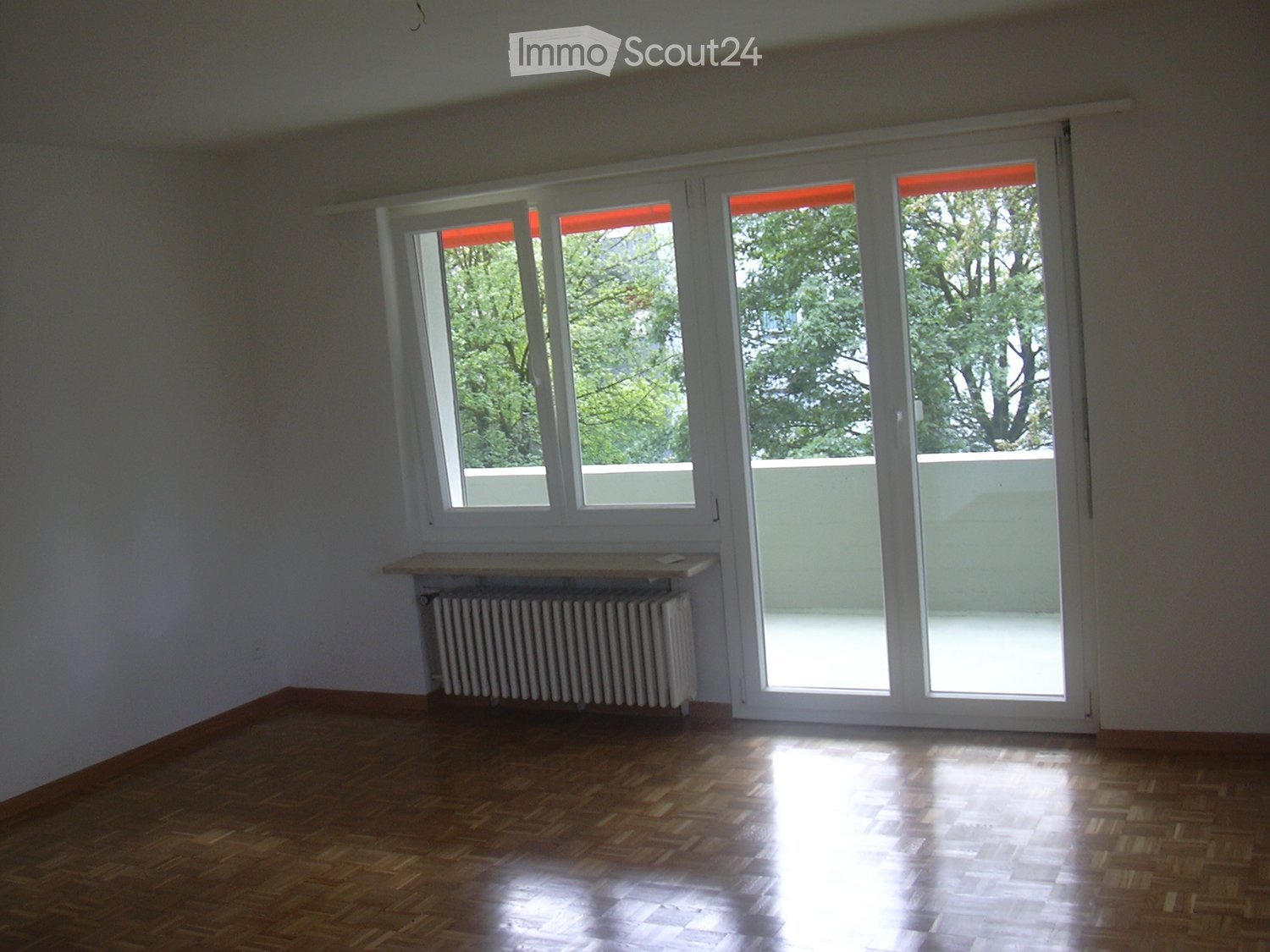 Empty room with wooden flooring, white walls, large windows, and a radiator under one window.