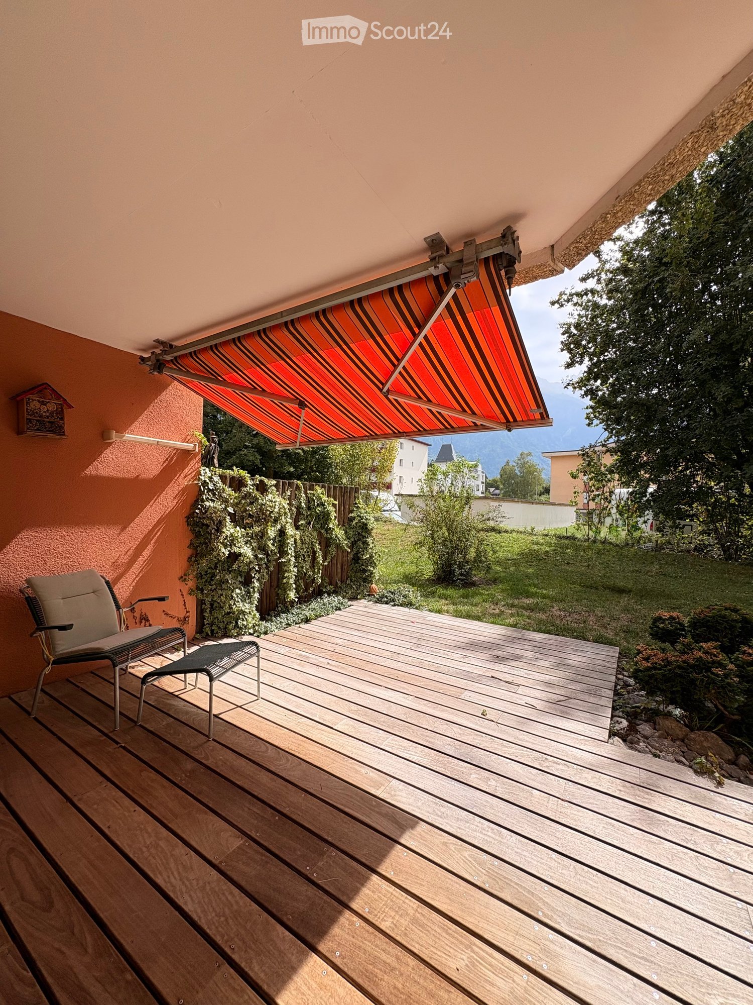 Wooden deck with a lounge chair and a small stool, featuring a retractable awning and a view of a green garden and mountains.