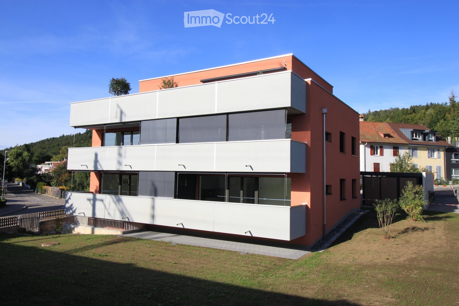 Modern two-story building with a combination of red and white exterior walls, large windows, and a flat roof. The building features a raised ground floor and is surrounded by a grassy area with a small fence and trees in the background.