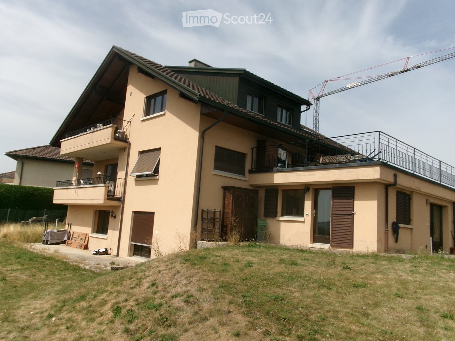 Two-story house with beige exterior, multiple windows, balconies, and a flat roof terrace. The house has a modern design with a mix of brown and green accents.