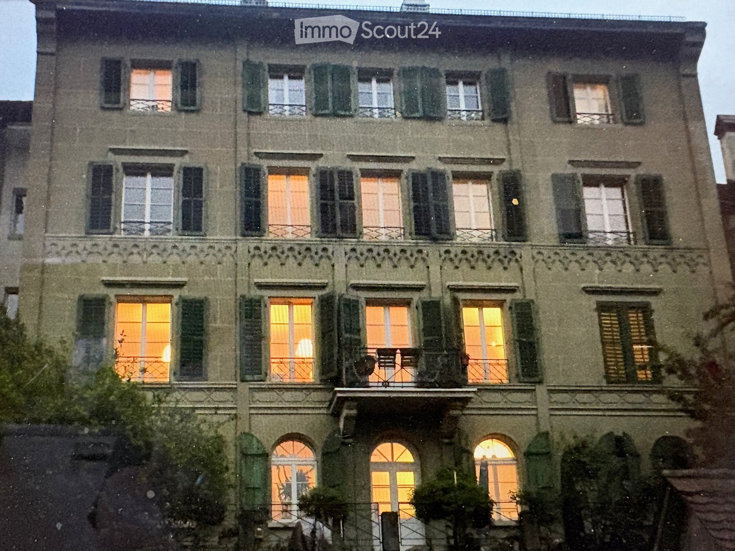 A multi-story residential building with multiple windows, some illuminated from within, and decorative balconies. The facade is made of stone with green shutters on the windows.
