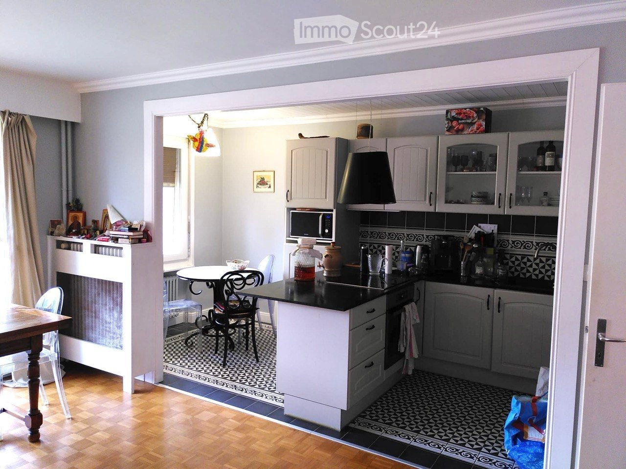 The image shows a modern kitchen with white cabinets, a black countertop, and a central island. There is a dining area with a round table and chairs, and a living area with a fireplace and a wooden table. The kitchen features various appliances, including