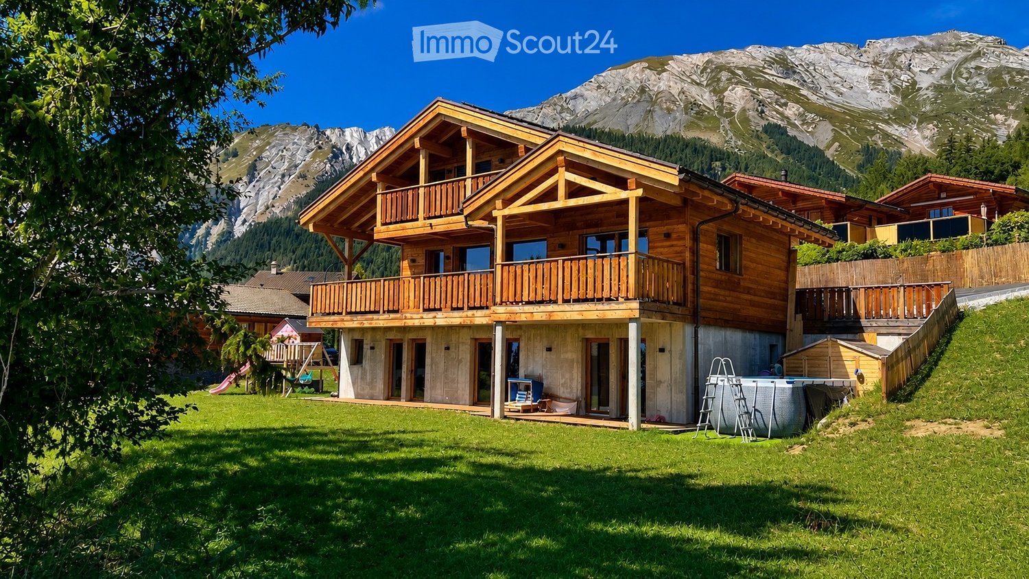A two-story wooden house with balconies, situated in a lush green yard with a view of snow-capped mountains in the background.