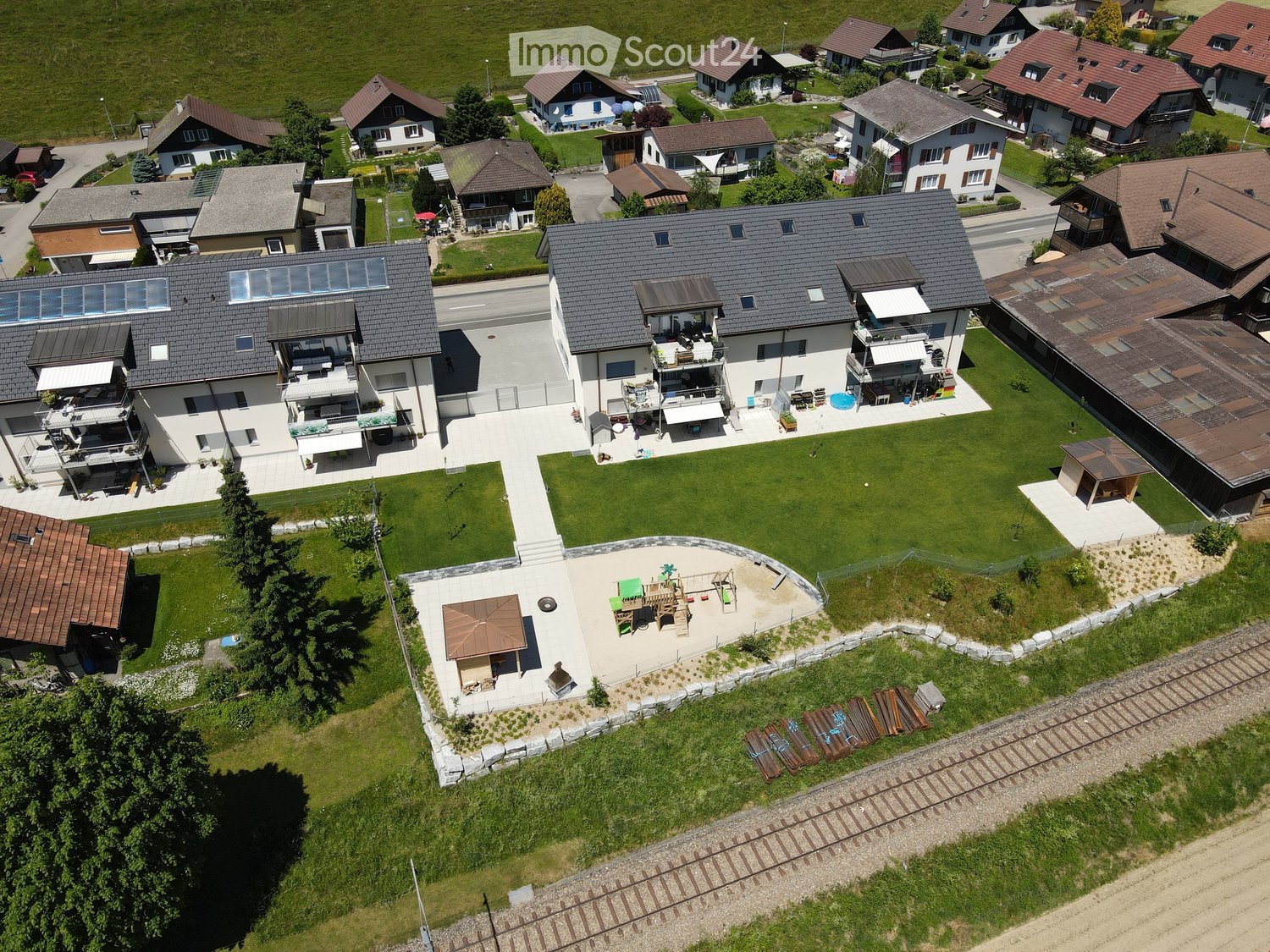 Aerial view of a residential area with multiple houses, green lawns, a playground, patio furniture, and a small pool.