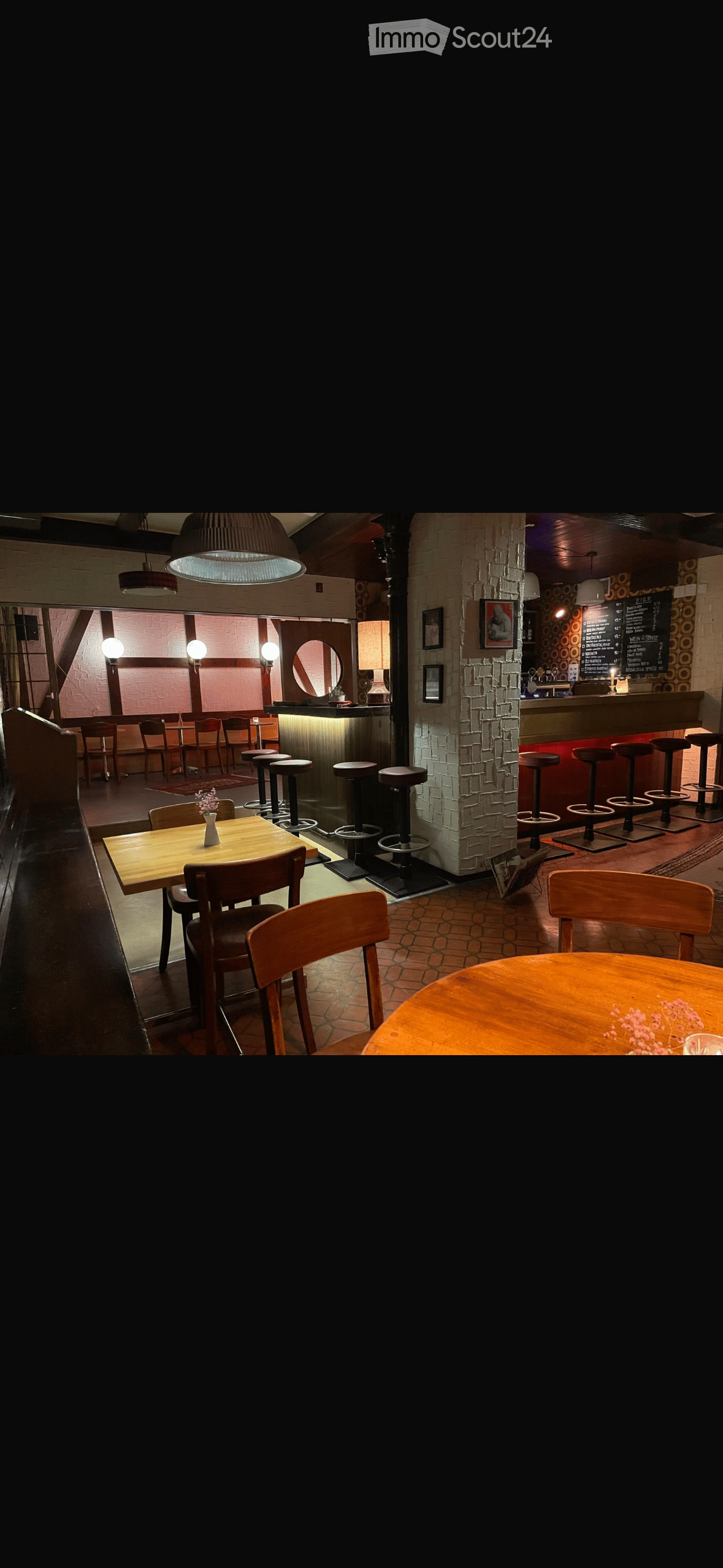 Cozy restaurant interior with wooden tables, chairs, bar stools, pendant lights, and a bar area with a menu board.