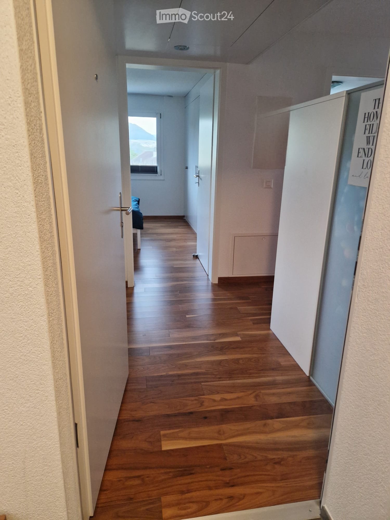 A long, narrow hallway with wooden flooring and white walls, featuring several doors and a visible glimpse of a room with a bed and a window.