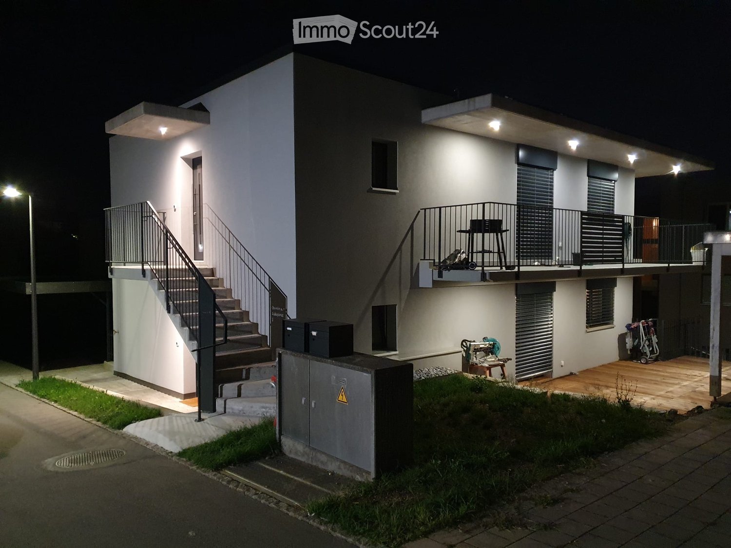 Modern two-story house with white walls, black shutters, and a raised ground floor. Features include a staircase leading to the upper level, a balcony with a railing, and outdoor storage items.