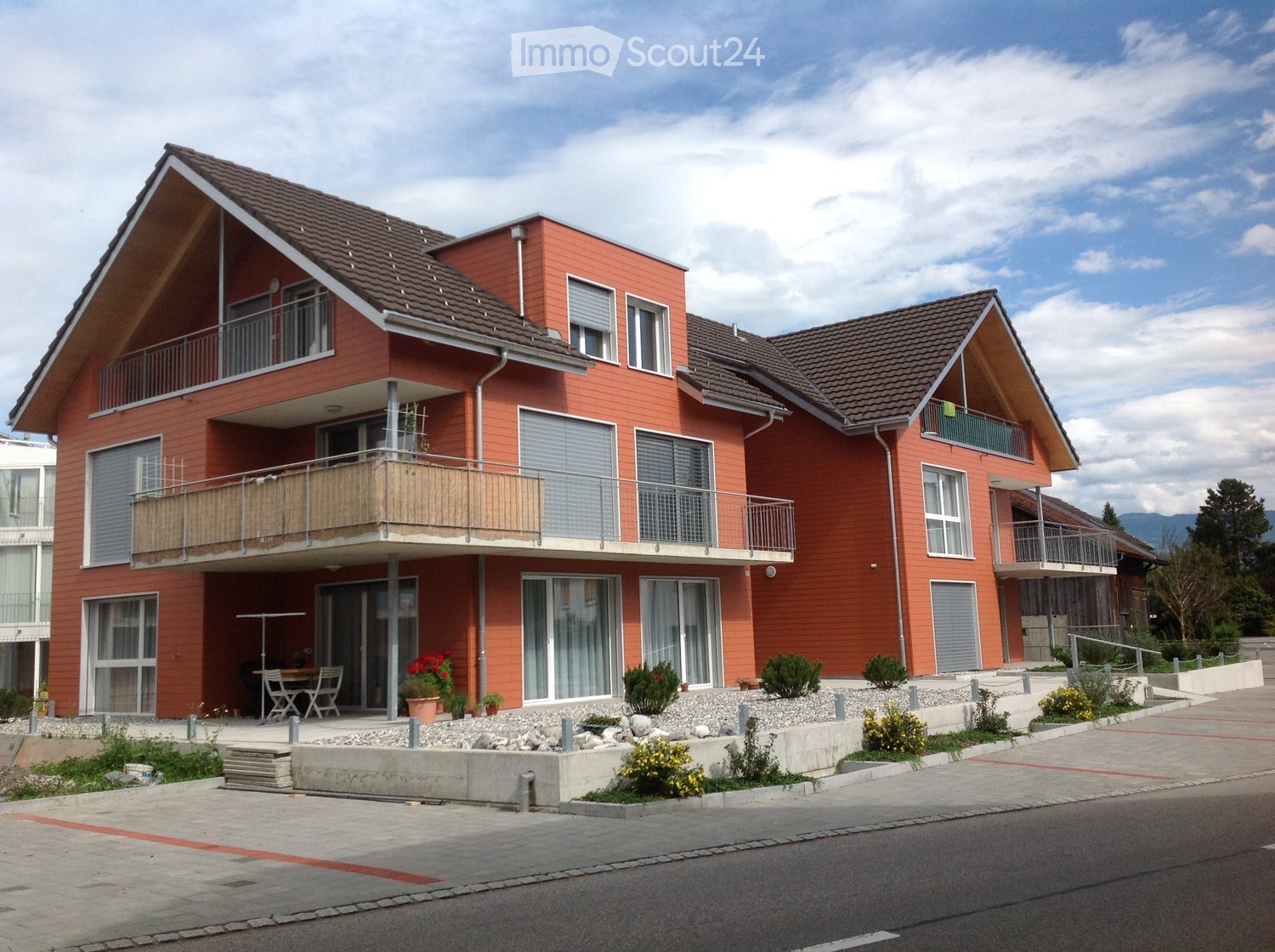 Red-colored exterior walls, brown roofs, glass doors and windows, balconies on each floor, shrubs and plants, table and chairs on ground floor balcony, parking area, road