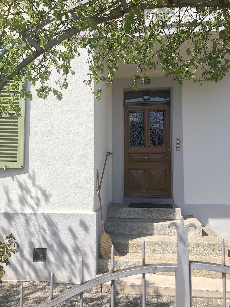 Entrance to a house with a wooden double door featuring decorative glass panels, flanked by white walls and a small staircase leading up to it. A broom leans against the wall beside the steps, and a metal gate is visible in the foreground.