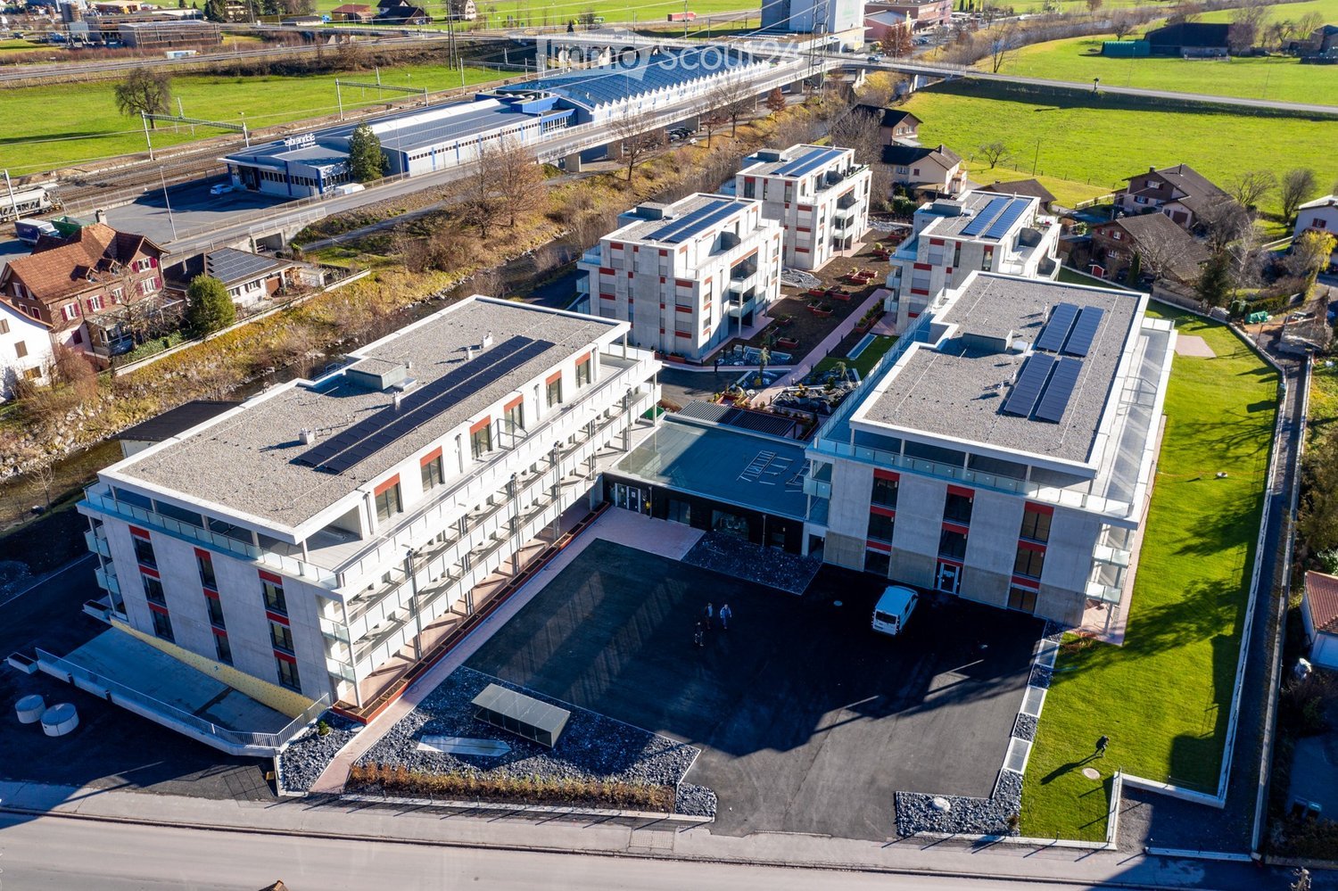 Group of residential buildings with modern architecture, multiple levels, solar panels on roofs, large parking area, trees, and green grass around.