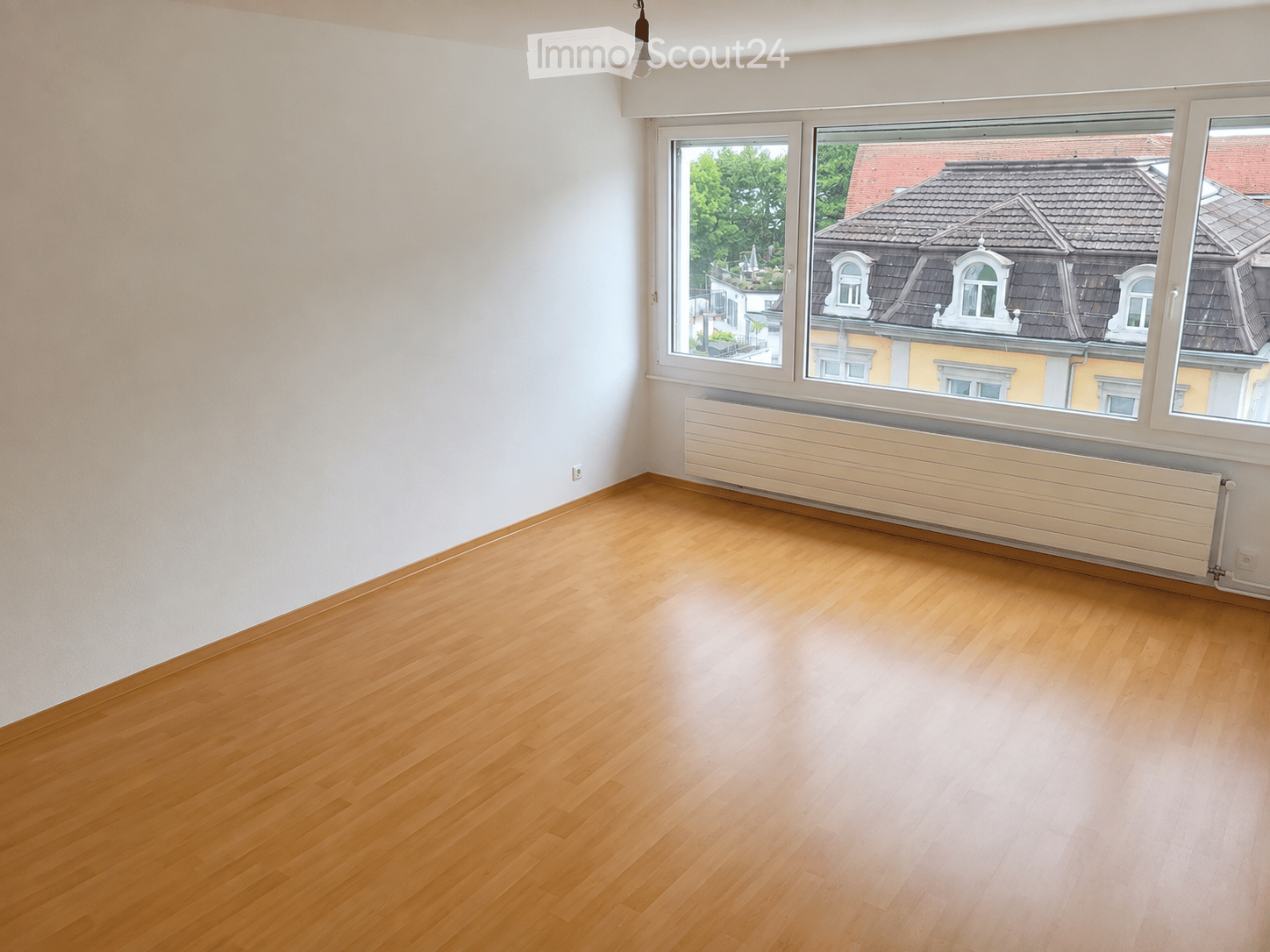 Empty room with wooden flooring, white walls, large windows, and a radiator under the windows.