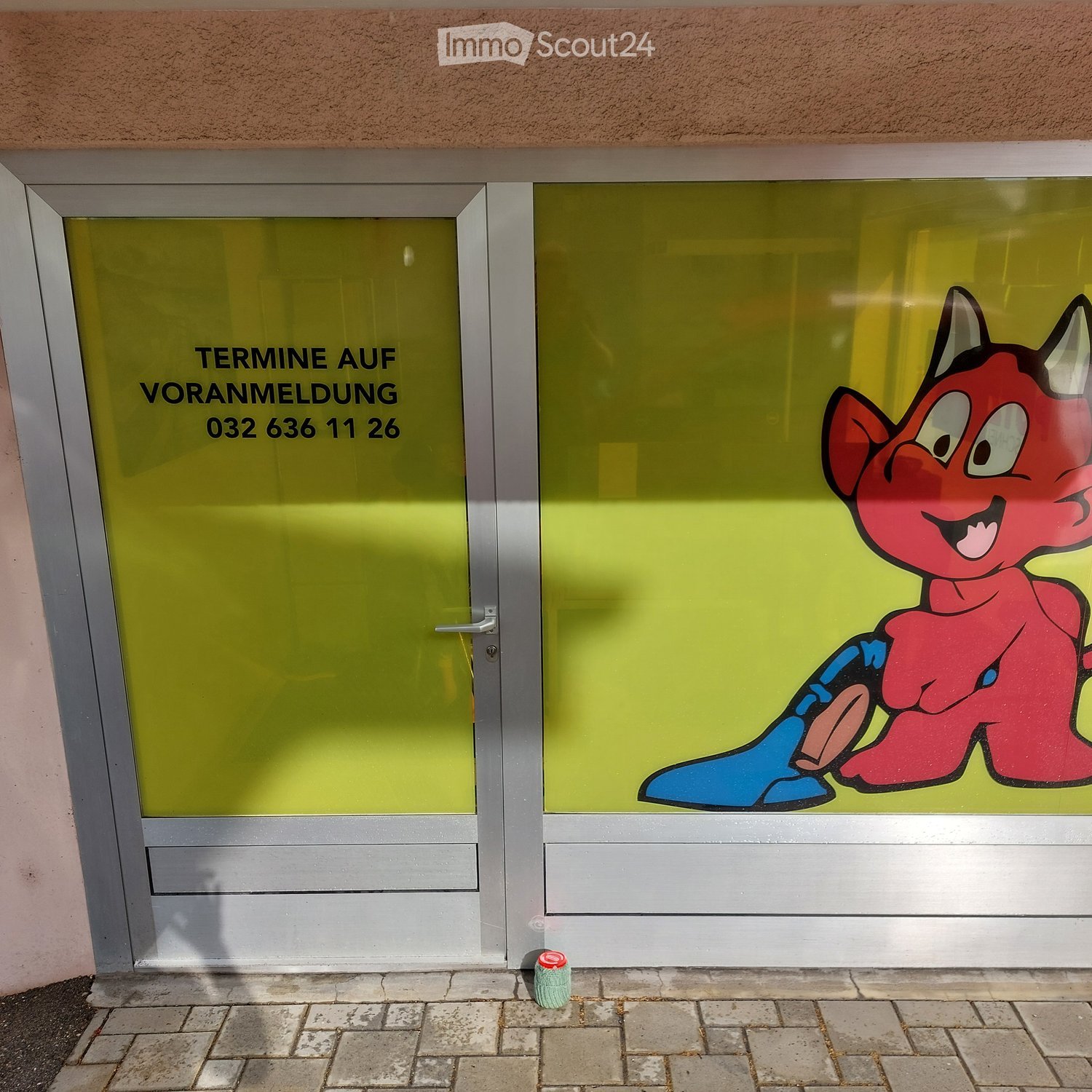 Building entrance with glass doors, green panels, and a cartoon character graphic on the right door. Text on the left door reads 'TERMINE AUF VORANMELDUNG 032 636 111 26'.