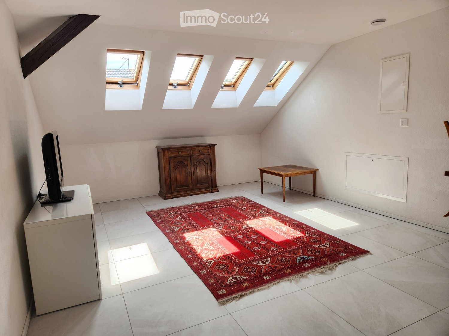 Bright attic room with white tiled flooring, wooden furniture including a cabinet and small table, a red patterned rug, and multiple skylights allowing natural light.