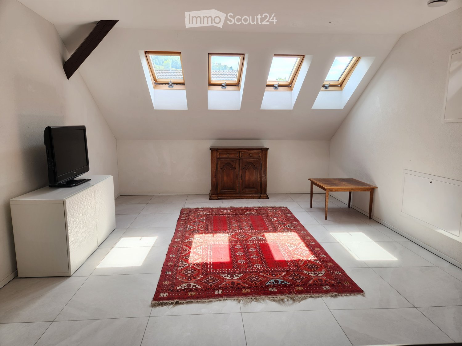 Bright attic room with skylights, tiled floor, red patterned rug, wooden cabinet, small table, and TV on a stand.
