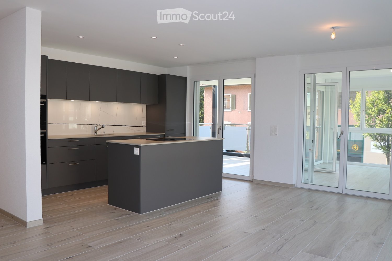 Modern kitchen with dark grey cabinets, a central island, and light wooden flooring. Large glass doors lead to an outdoor area.