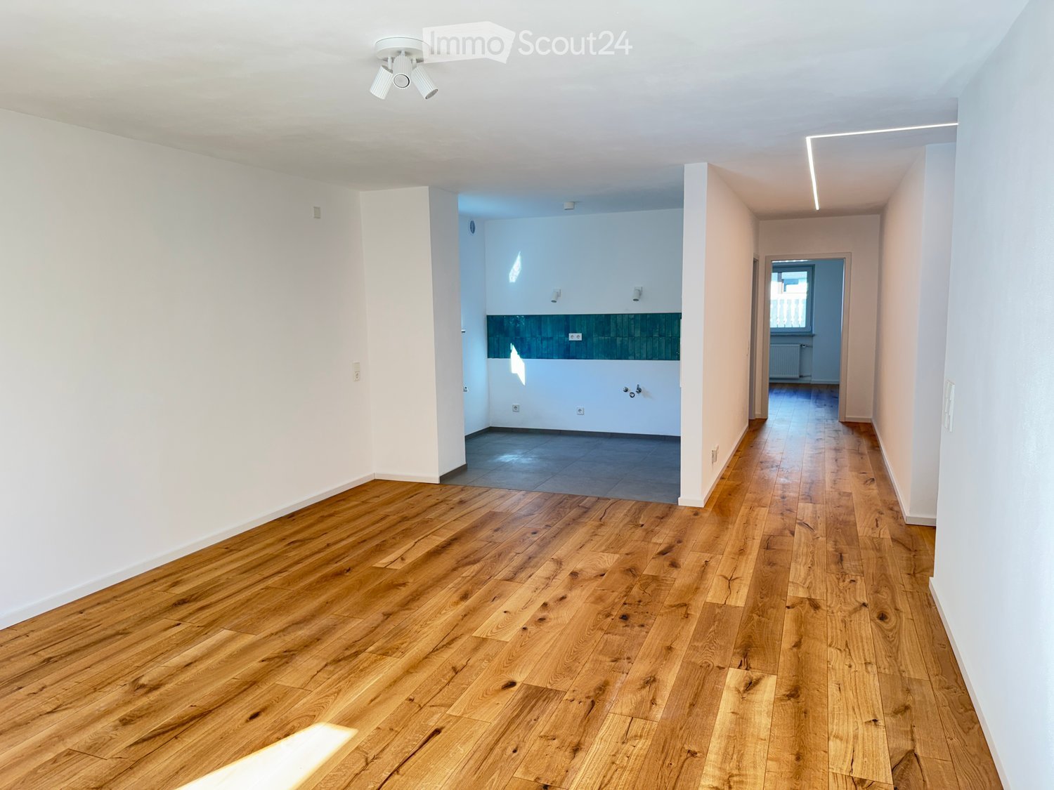 Empty hallway with wooden flooring, white walls, and recessed ceiling lighting. The hallway leads to a tiled room and a windowed area with a radiator.