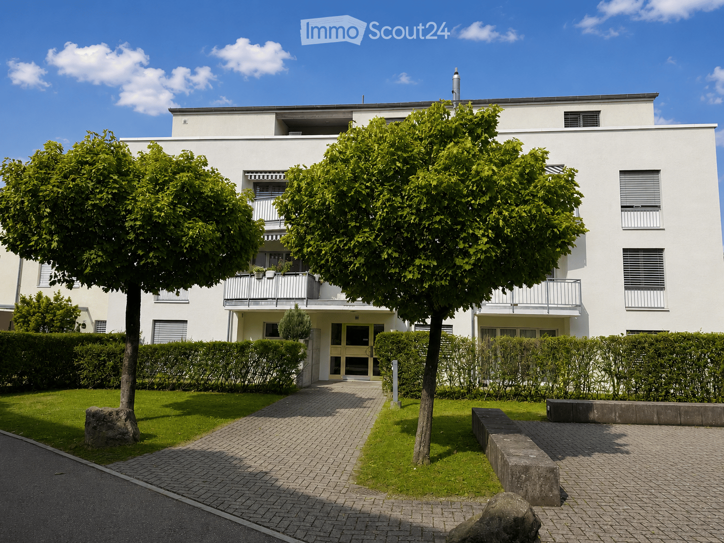 Modern two-story white building with balconies, surrounded by greenery and a paved walkway.