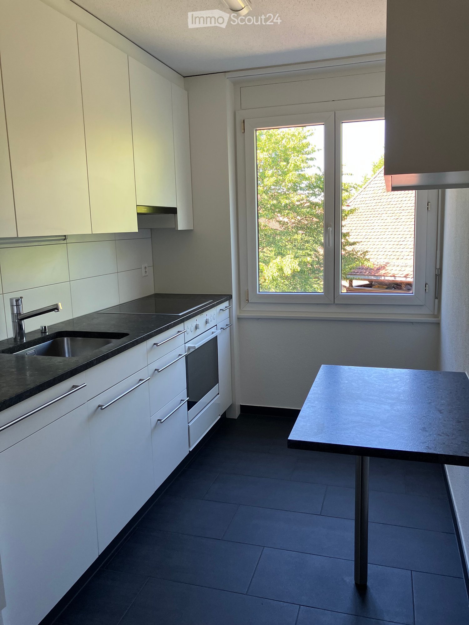 Modern kitchen with white cabinets, black countertops, stainless steel sink and faucet, built-in oven, and a small black dining table.